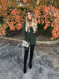 smiling woman in green sweater and tall black boots under autumn trees