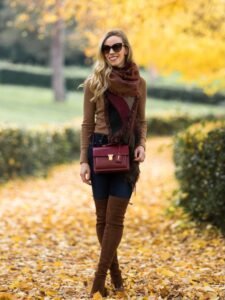 woman standing on autumn leaves wearing a brown scarf, burgundy handbag, and tall brown boots