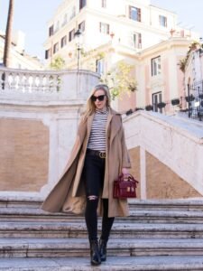 woman in camel coat and striped turtleneck walking down stone steps holding a red handbag