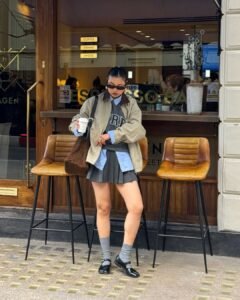 woman in a tan barn jacket and pleated mini skirt standing outside a cafe