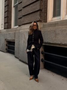 woman in black napoleon jacket and dark barrel jeans standing against a historic stone building.