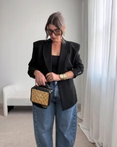 woman in black blazer and blue barrel jeans holding a gold and black handbag indoors.