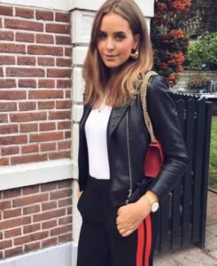 woman in black leather jacket and white top standing against a brick wall with red bag