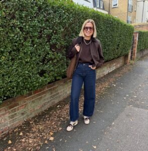 woman in brown jacket and dark barrel jeans standing on sidewalk next to green hedge.
