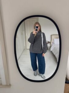 woman taking mirror selfie wearing grey sweater and blue leopard print barrel jeans in bedroom.