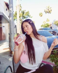 woman with long hair drinking a smoothie outdoors wearing a white top and pink bag