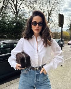 woman wearing a white button-down shirt and blue jeans holding a brown suede bag
