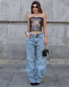 woman in metallic tube top and light wash barrel jeans standing against stone wall.