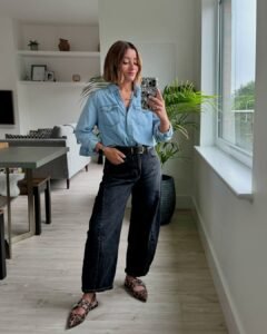 woman in light denim shirt and black barrel jeans holding phone in a bright room.