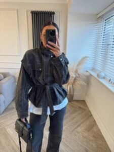 woman wearing belted dark grey jacket and black jeans taking mirror selfie