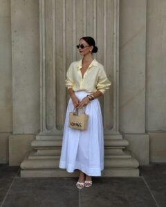 woman wearing a pale yellow button-down shirt and white maxi skirt with a straw bag.