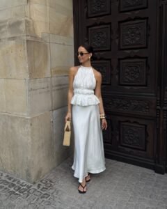 woman in a white halter top and satin maxi skirt standing by a wooden door.