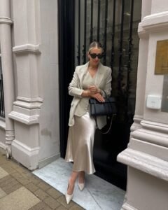woman in cream blazer and satin midi skirt standing by black door