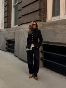 woman in black napoleon jacket and dark barrel jeans standing against a historic stone building.