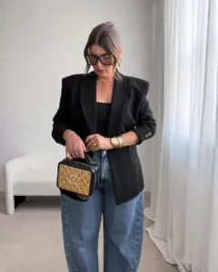 woman in black blazer and blue barrel jeans holding a gold and black handbag indoors.