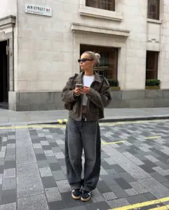 woman in brown bomber jacket and charcoal barrel jeans standing on a city street corner.