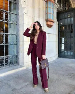 woman wearing a deep burgundy suit and tan turtleneck holding a patterned designer handbag.