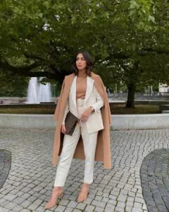 woman wearing a cream patterned suit and tan overcoat standing near a park fountain.