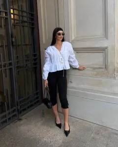 woman wearing a white ruffled blouse, black capri pants, and pointed-toe heels standing outdoors.