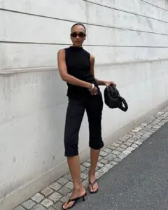 woman wearing a black sleeveless turtleneck and black capri pants with a woven handbag.