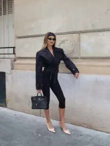 woman wearing a cropped black trench coat and black capri pants holding a leather bag.