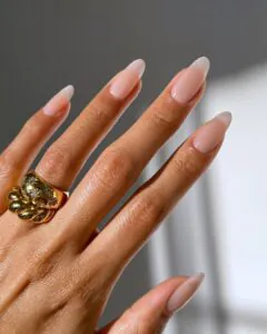 sheer neutral lip gloss nails on long almond shape with chunky gold statement ring.