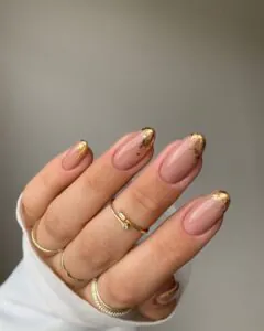 nude almond nails with distressed gold foil tips and thin gold stacking rings.