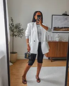 woman wearing a white blazer, striped top, and black capri pants holding a small handbag.