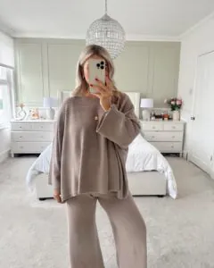 woman in a beige oversized sweater and matching wide-leg trousers standing in a white bedroom.