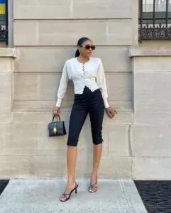 woman in a white button-up cardigan and black capri pants posing against a stone building.