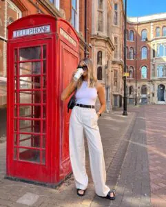 blonde woman in white trousers drinking coffee next to a red london telephone booth