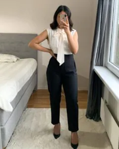 woman wearing a white sleeveless bow blouse and high-waisted black trousers in a bedroom.