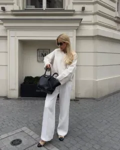 blonde woman in an ivory sweater and white wide-leg trousers standing on a city sidewalk.