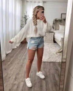mirror selfie of woman with long braid wearing white knit sweater and distressed denim shorts.