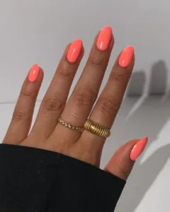 vibrant coral orange almond nails with two gold rings on tanned skin