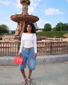 woman wearing white off-the-shoulder top and long denim shorts standing by a stone fountain.