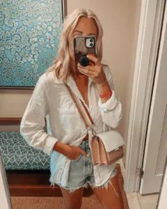 mirror selfie of woman wearing white button-down shirt and frayed hem denim shorts.