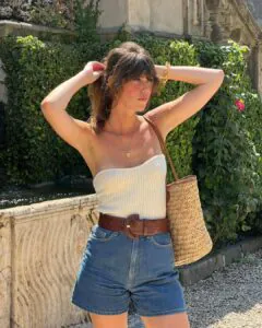 woman wearing white strapless top, denim shorts, and brown belt holding a straw bag.