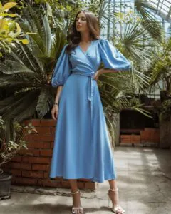 woman in blue silk wrap dress with puffy sleeves standing in a tropical greenhouse