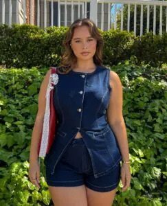 woman wearing matching dark wash denim vest and shorts set standing in front of greenery.