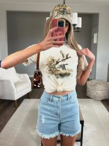 woman wearing cream graphic t-shirt, light blue denim shorts, and camouflage baseball cap.