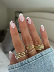 almond French tip nails with small strawberry art accents and gold stacked rings