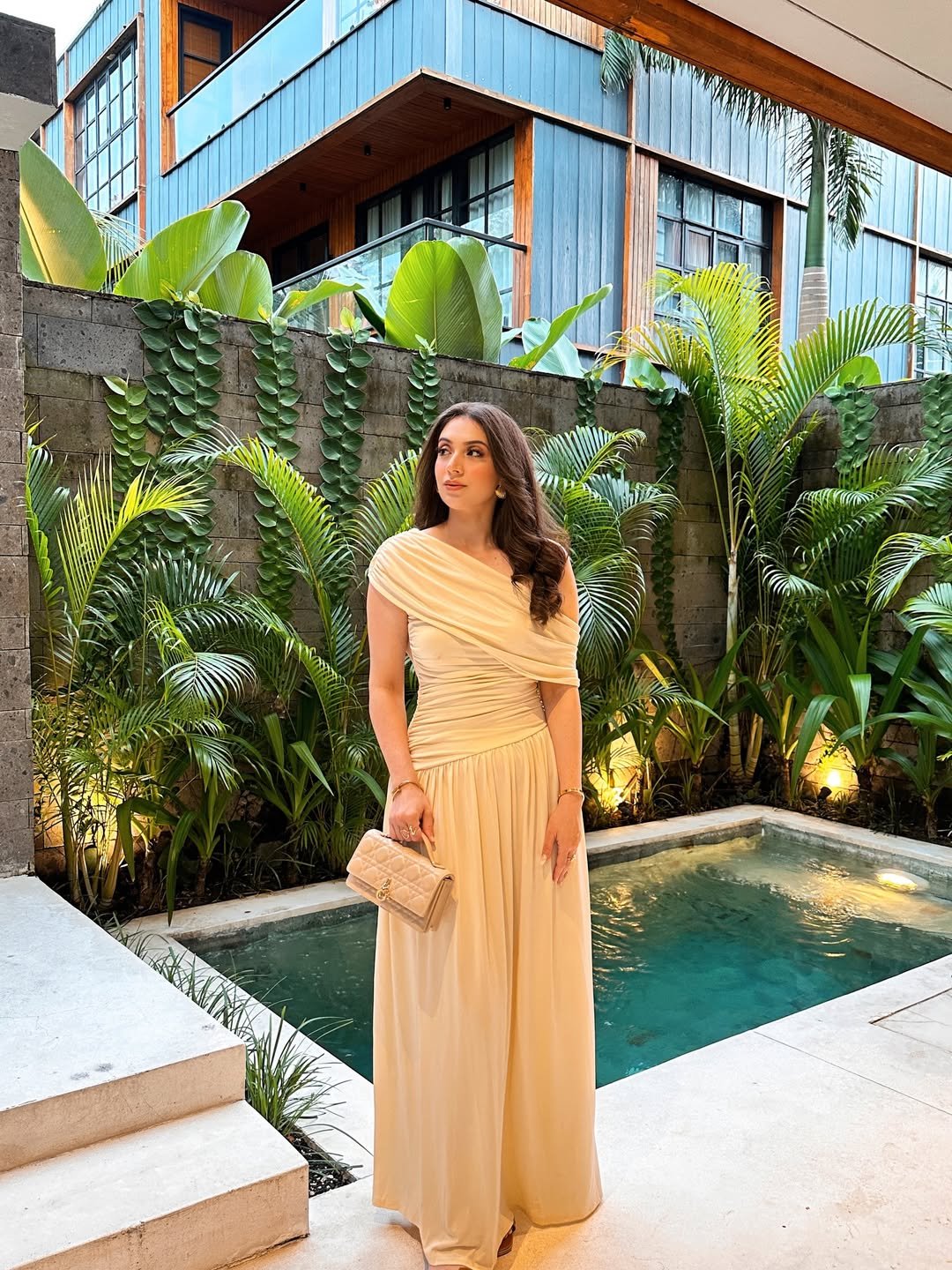 woman in cream off-the-shoulder maxi dress standing by tropical pool with lush greenery