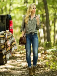 blonde woman in green button down shirt and blue jeans standing near a forest trail