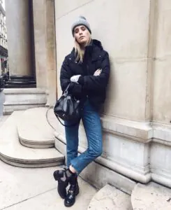 woman in black puffer jacket and blue jeans leaning against a stone building in winter