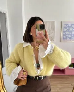 woman in a yellow mohair cardigan over a white ruffled shirt taking a mirror selfie