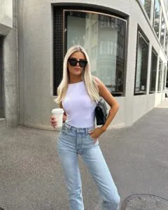 woman wearing sleeveless white bodysuit and light blue jeans while holding coffee cup outdoors.