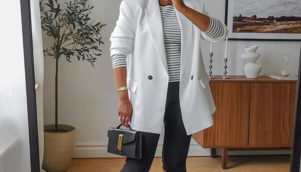 woman wearing a white blazer, striped top, and black capri pants holding a small handbag.