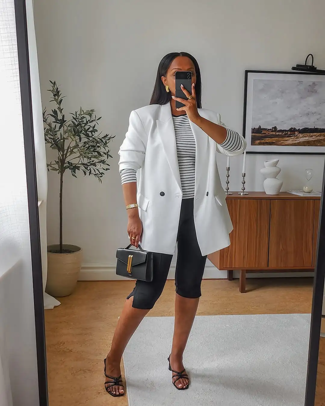 woman wearing a white blazer, striped top, and black capri pants holding a small handbag.