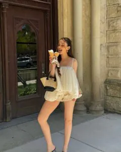 woman in a cream-colored ruffled top holding two ice cream cones near a stone building.